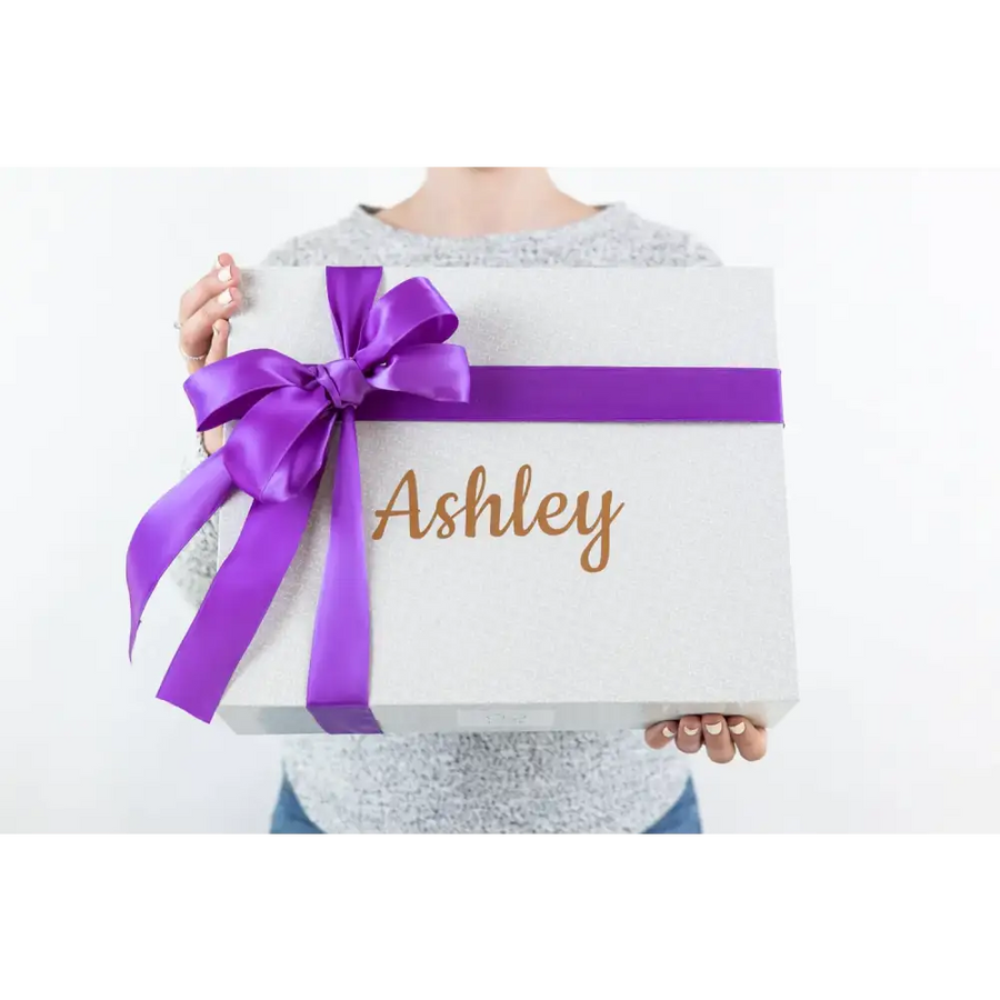 A white glittery gift box adorned with a vibrant purple satin ribbon bow and the name ’ashley’ in gold cursive lettering.