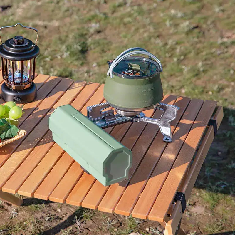 Olive green camping kettle with a silver handle and lid, resting on a portable stove atop a wooden table