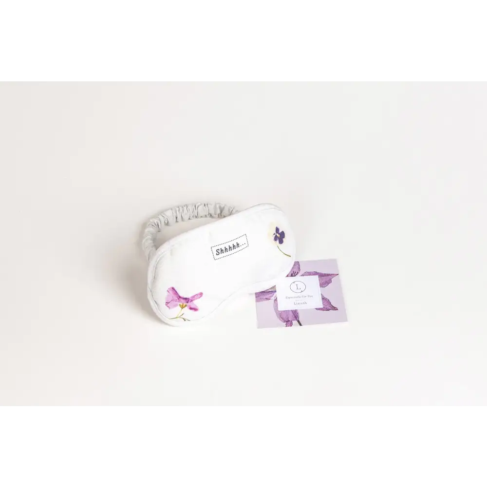 White sleep mask with delicate purple floral embroidery and a soft elastic strap, featuring the word ’shhh...’ stitched on its surface.