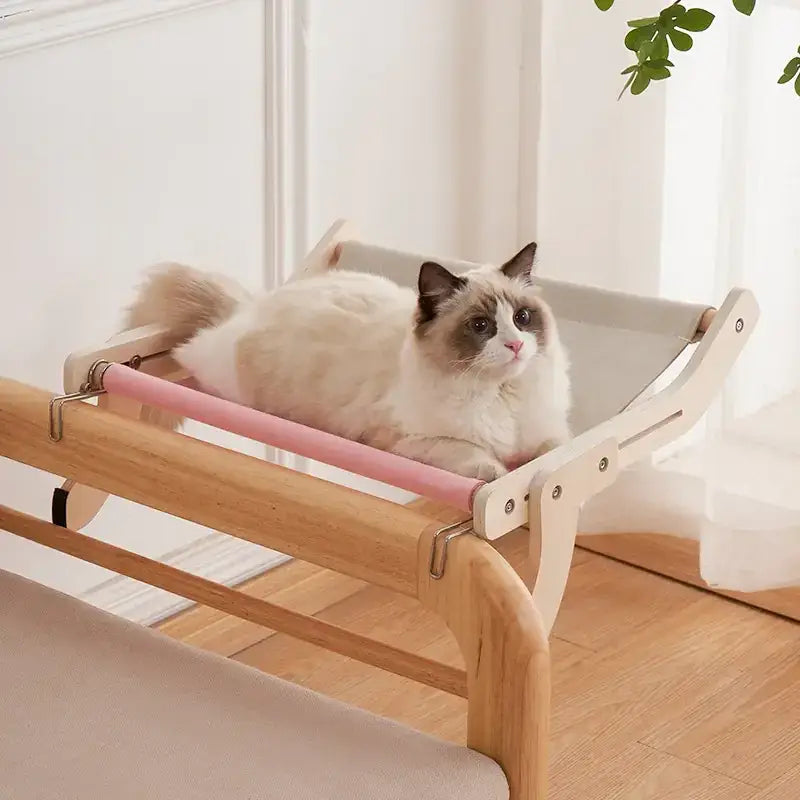A cozy cat hammock with a light wood frame and soft pink fabric straps, featuring a plush beige cushion for comfort.