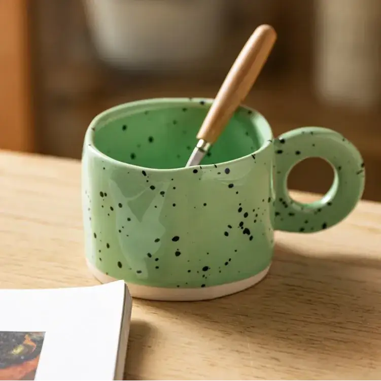 A mint green ceramic mug with black speckles and a circular handle, holding a wooden-handled spoon.