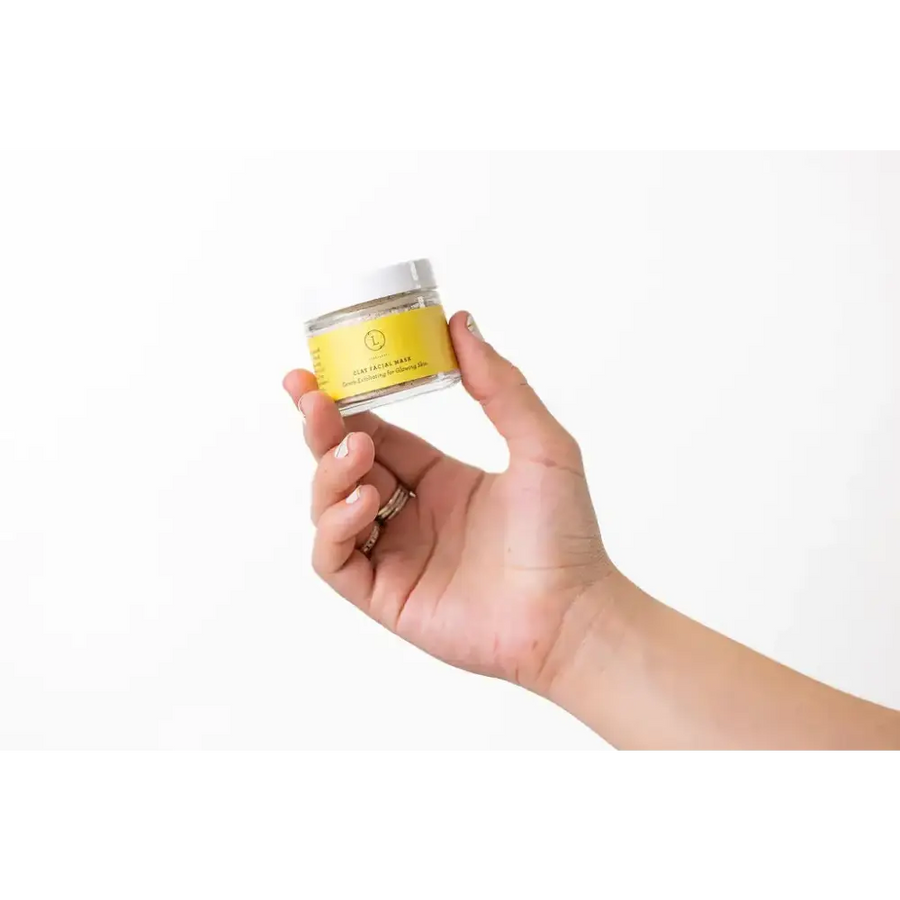 A clear glass jar with a white lid and a bright yellow label, containing a clay facial mask.