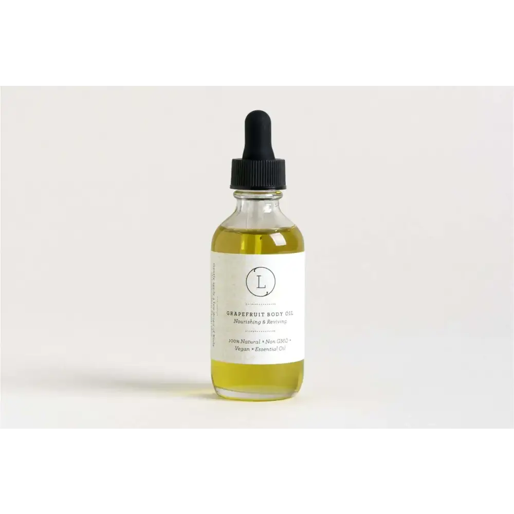 A clear glass bottle with a black dropper cap contains golden-yellow grapefruit body oil, labeled as 100% natural and vegan.