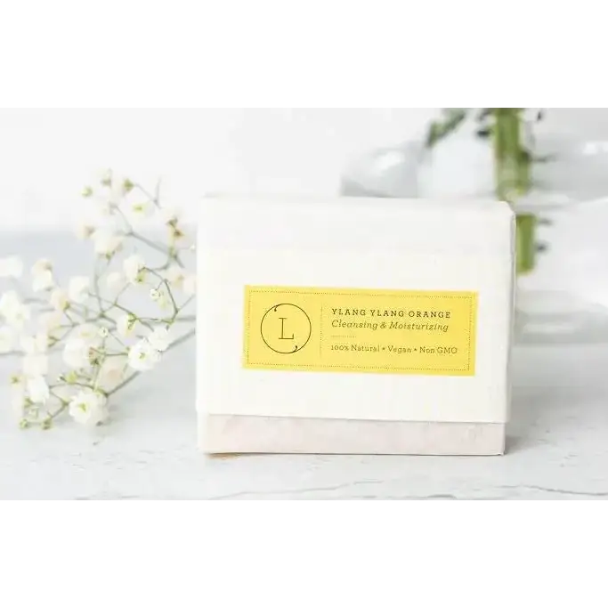A white rectangular bar of soap with a yellow label that reads ’ylang ylang orange cleansing & moisturizing’ and features a circular logo.