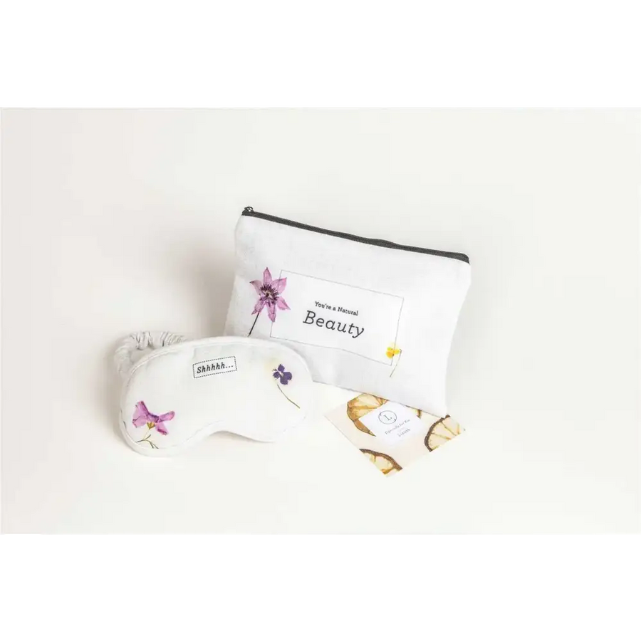 White eye mask with delicate purple floral embroidery and a small ’shhh...’ label, paired with a matching white pouch featuring the phrase “you’re a natural beauty” in black text.