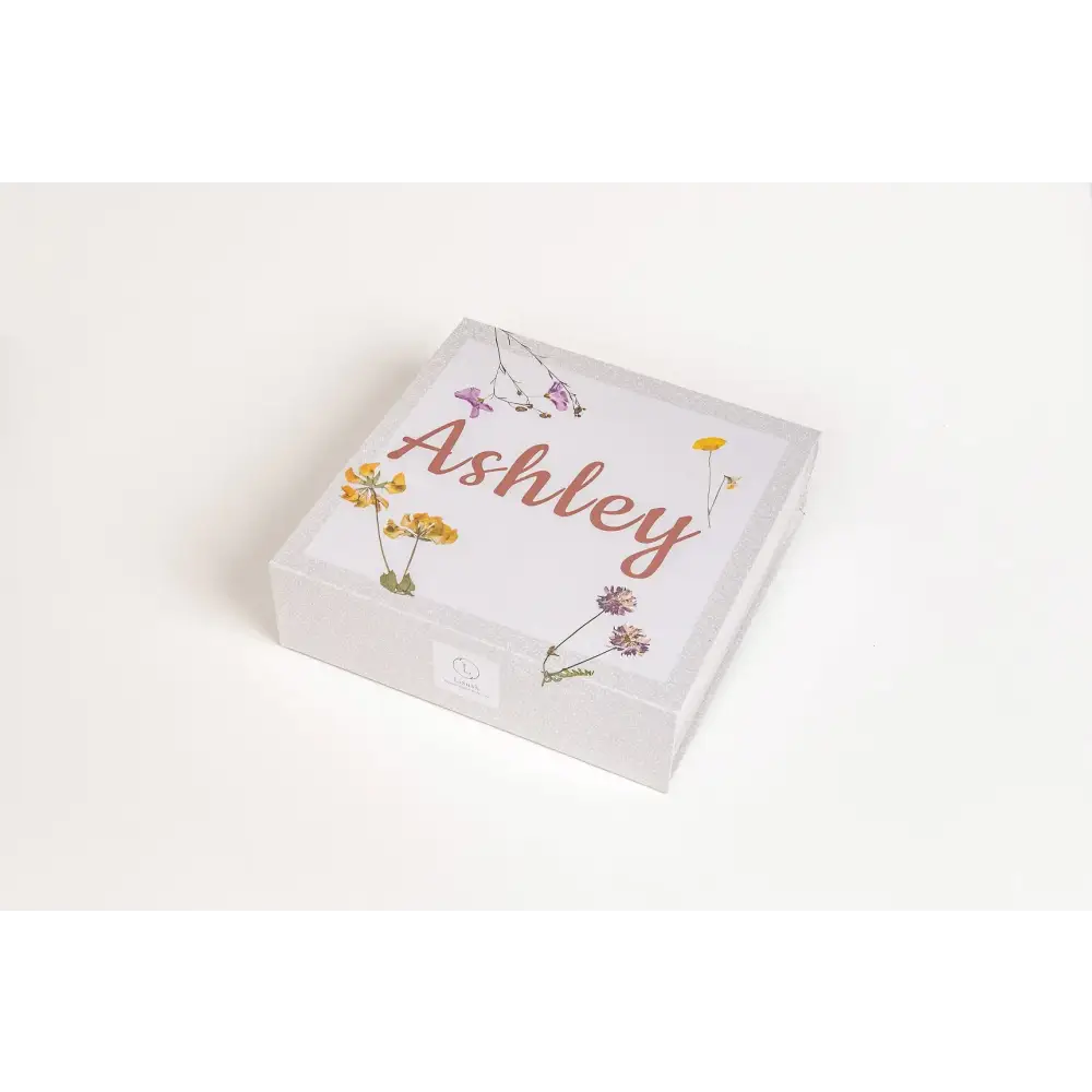 A white square gift box adorned with delicate pressed flowers in yellow, purple, and pink hues around the name ’ashley’ written in a warm terracotta cursive script.