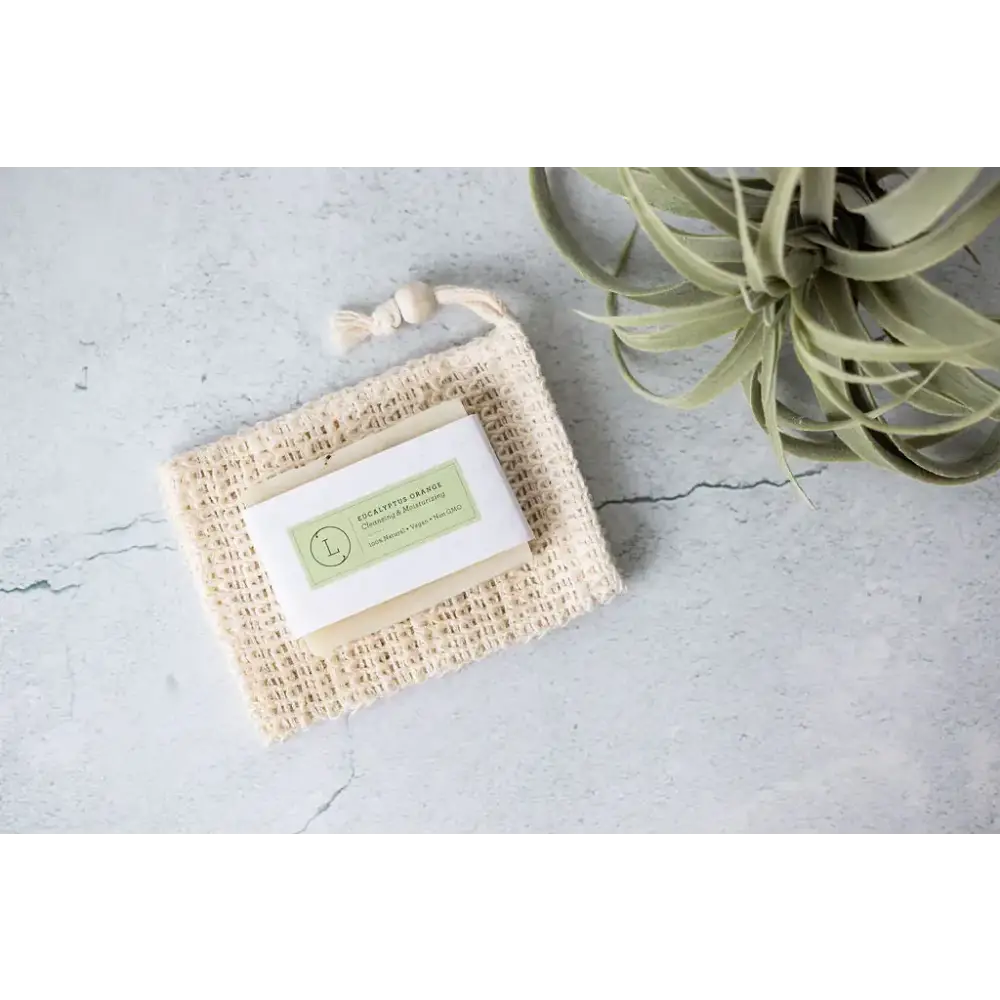 Beige woven mesh soap saver with a white label featuring green text and logo, holding an eucalyptus orange bar of soap.