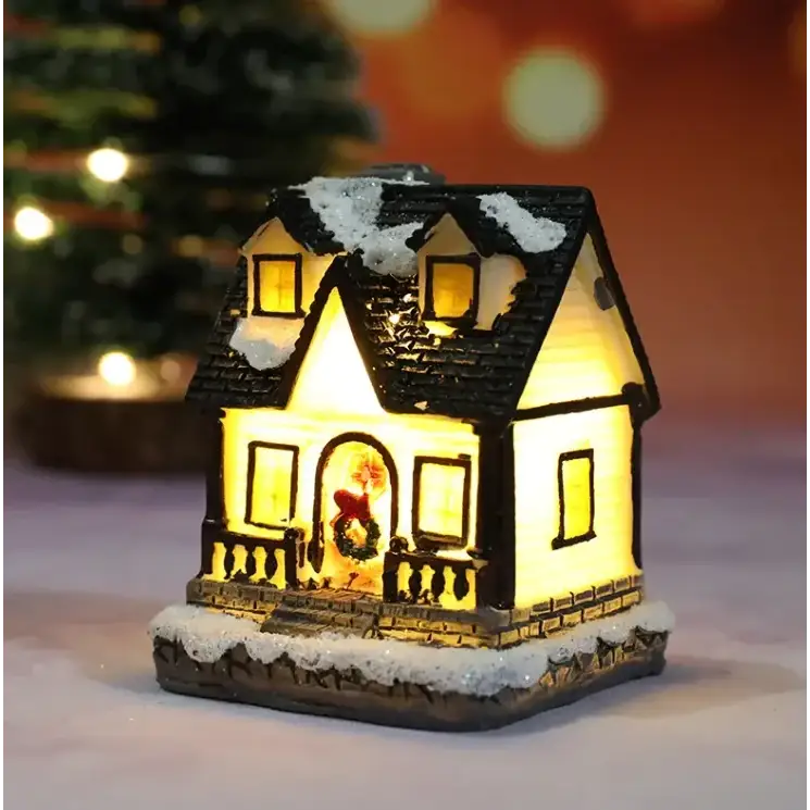 A warmly lit miniature christmas house figurine with a black shingled roof dusted in white snow, glowing yellow interior lights, and a festive wreath adorning the front door.