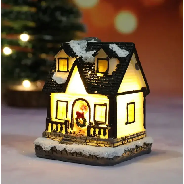 A warmly lit miniature christmas house figurine with a black shingled roof dusted in white snow, glowing yellow interior lights, and a festive wreath adorning the front door.