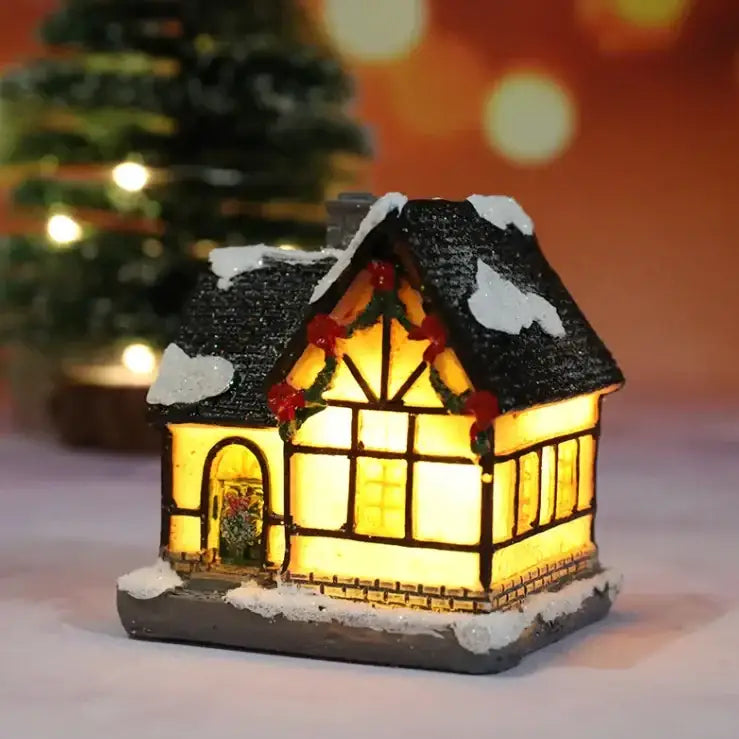 A miniature illuminated cottage with a black shingled roof dusted in white snow, glowing yellow windows framed by dark timber beams and adorned with red berries on green garlands.