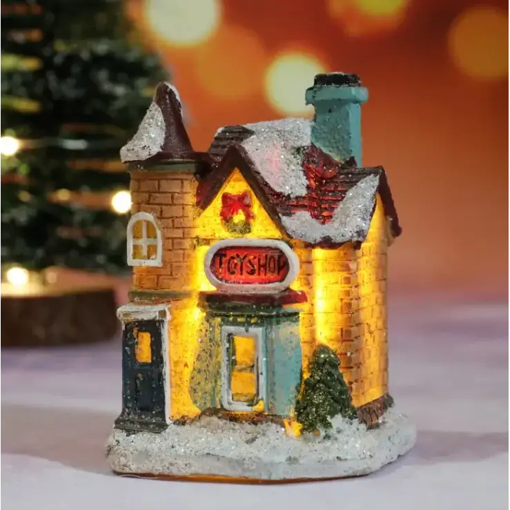 A miniature illuminated toy shop figurine with a brick facade, snow-dusted roof and chimney, glowing yellow windows, and an oval red sign reading ’toyshop.’