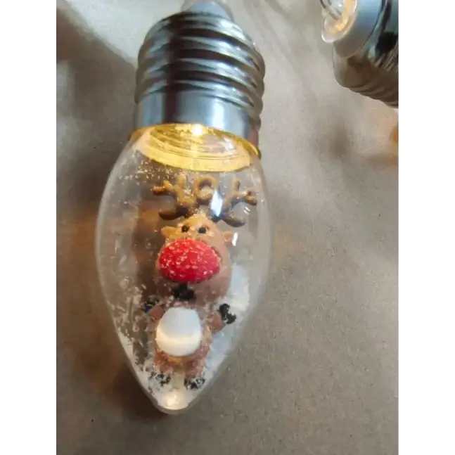 A clear glass christmas light bulb featuring a festive reindeer figurine with antlers, a red nose, and white belly surrounded by faux snow.