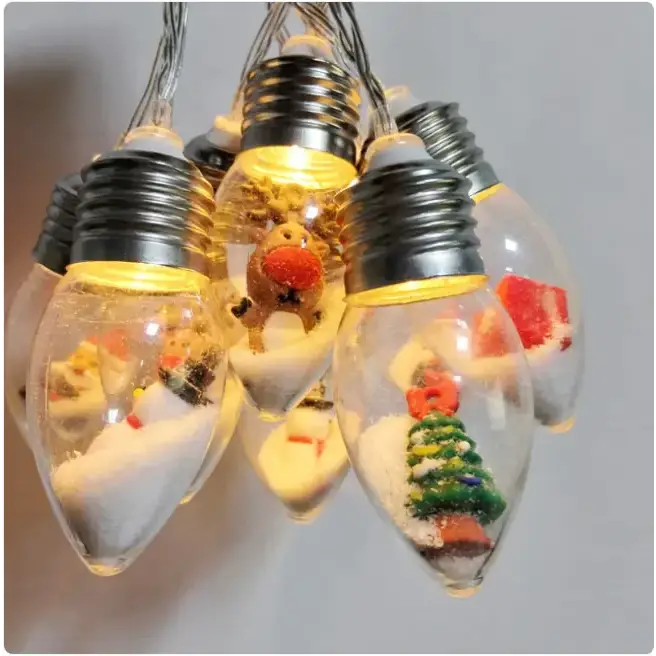 String of clear glass christmas light bulbs, each containing miniature festive scenes with snow and figurines like reindeer and trees.