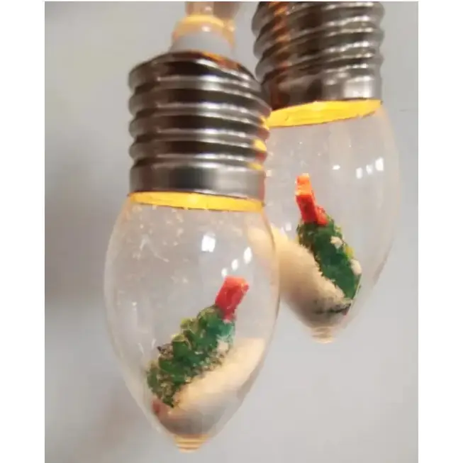 Clear glass light bulb ornaments with miniature green christmas trees and red toppers nestled in white sand.