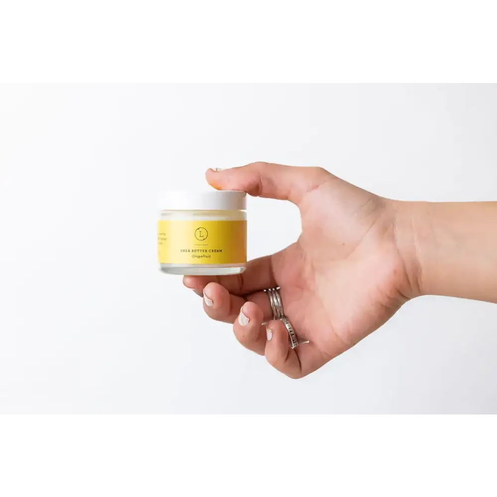 A small, clear glass jar with a white lid and a vibrant yellow label that reads ’shea butter cream’ by the brand lush.