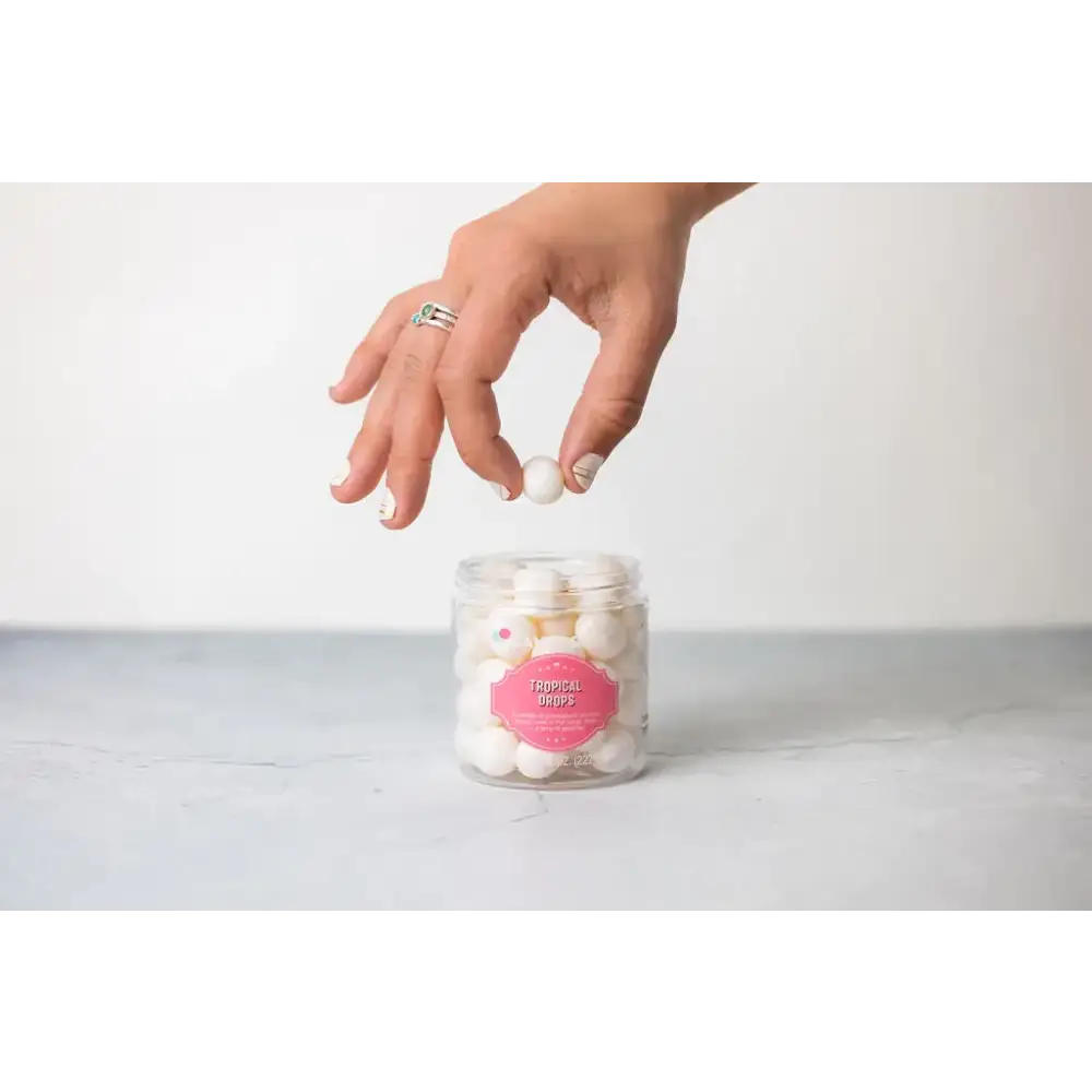 A hand reaches for a white, spherical candy from a clear glass jar labeled ’tropical drops.’