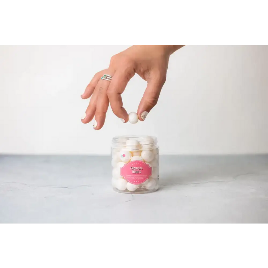 A hand reaches for a white, spherical candy from a clear glass jar labeled ’tropical drops.’