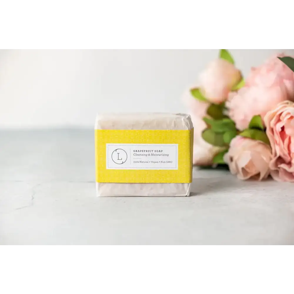 A rectangular bar of pale yellow grapefruit soap wrapped in a bright yellow label with black text and a circular logo, presented on a light surface beside soft pink flowers.