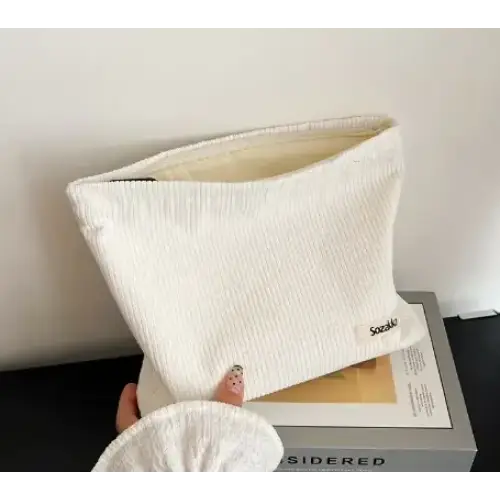 A cream-colored corduroy clutch with a subtle textured weave and a small embroidered brand tag on the corner.