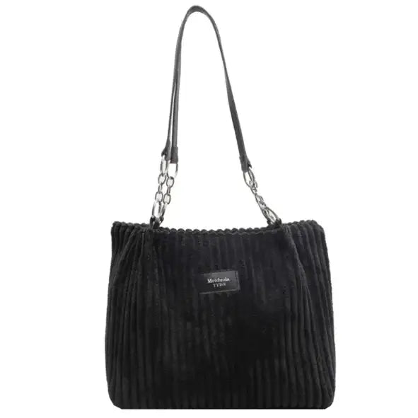 Black corduroy tote bag with a chain and leather strap, featuring a small rectangular brand label on the front.