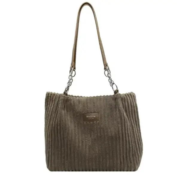 A taupe corduroy tote bag with a textured vertical ribbed pattern, featuring a brown leather strap attached by silver-toned metal rings and a small rectangular brand tag on the front.