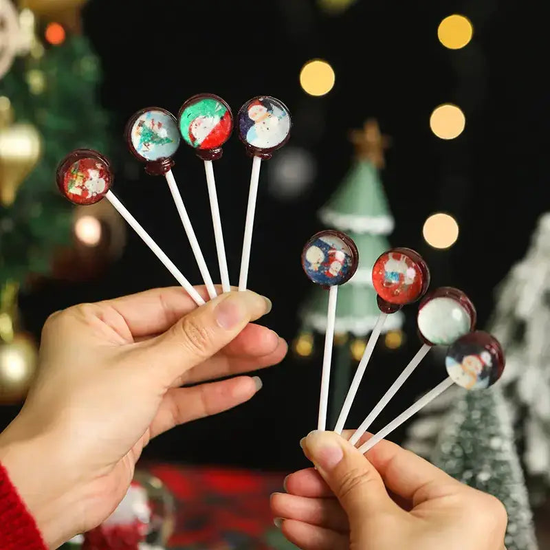 Christmas-themed lollipops with festive holiday designs on clear, glossy candy spheres mounted on white sticks.