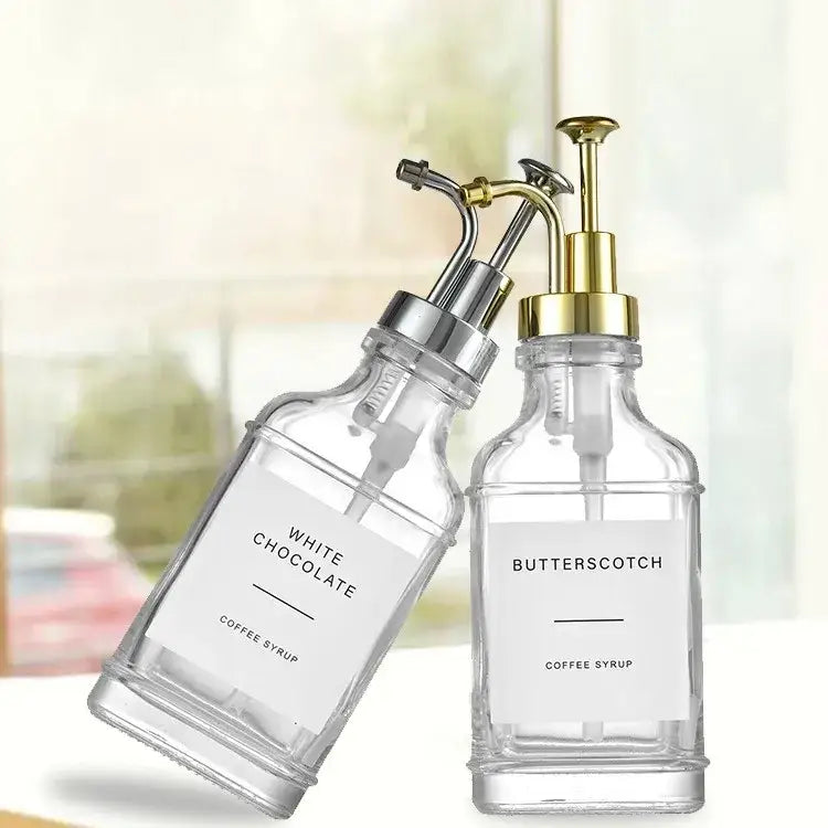 Clear glass syrup dispensers with gold and silver pump tops, labeled ’white chocolate’ and ’butterscotch coffee syrup.’