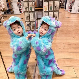 Two toddlers in matching fluffy blue and purple monster costumes with long tails, hooded heads featuring large white eyes.