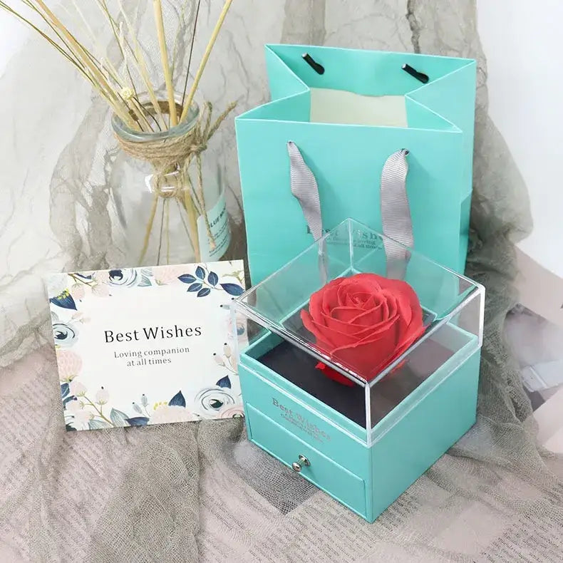 A vibrant red preserved rose nestled inside a clear acrylic box with a turquoise drawer and matching gift bag.