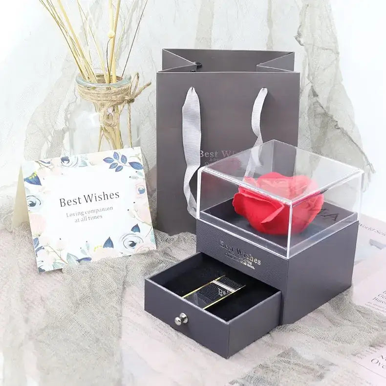 A vibrant red rose encased in a clear acrylic box atop a sleek gray gift set with an open drawer revealing a gold-trimmed lipstick.