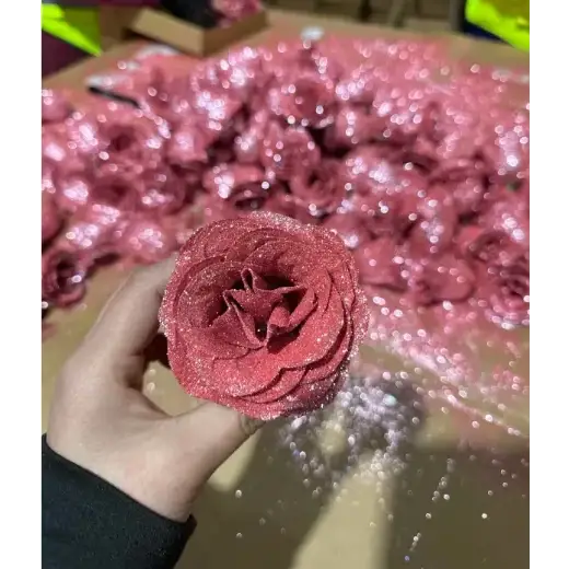 A hand holds a glittery pink fabric rose with layered petals and sparkling texture.