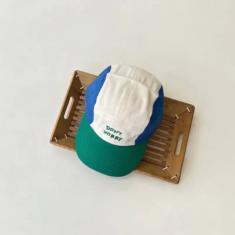 A colorful baseball cap with a white front panel, blue side panels, and a green brim, featuring the embroidered text ’don’t worry’ in green.
