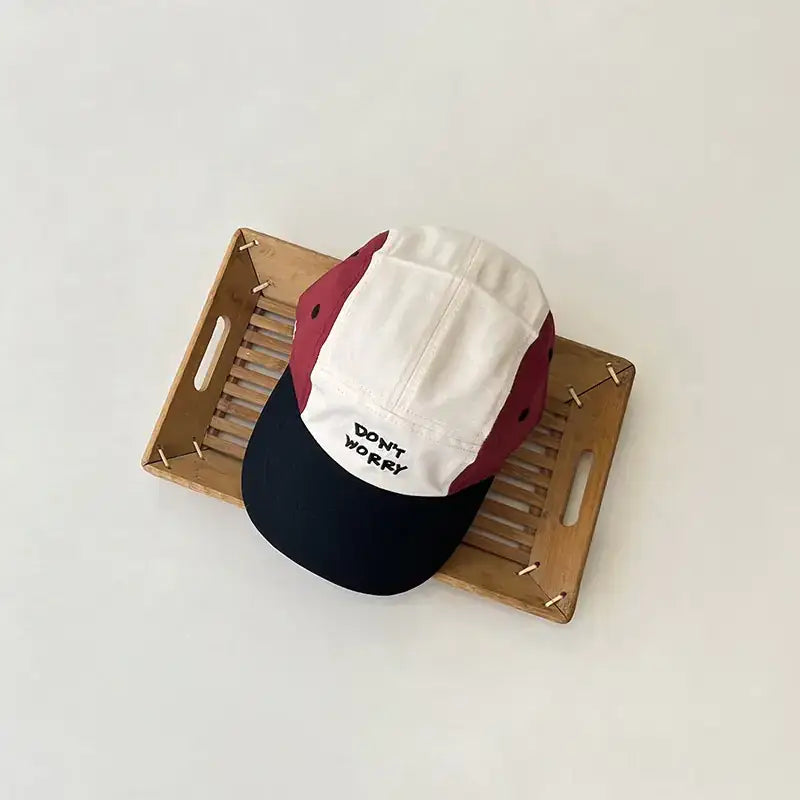 A stylish multi-colored baseball cap with a white front panel, maroon side panels, and a black brim, featuring the embroidered text ’don’t worry’ in bold black letters.
