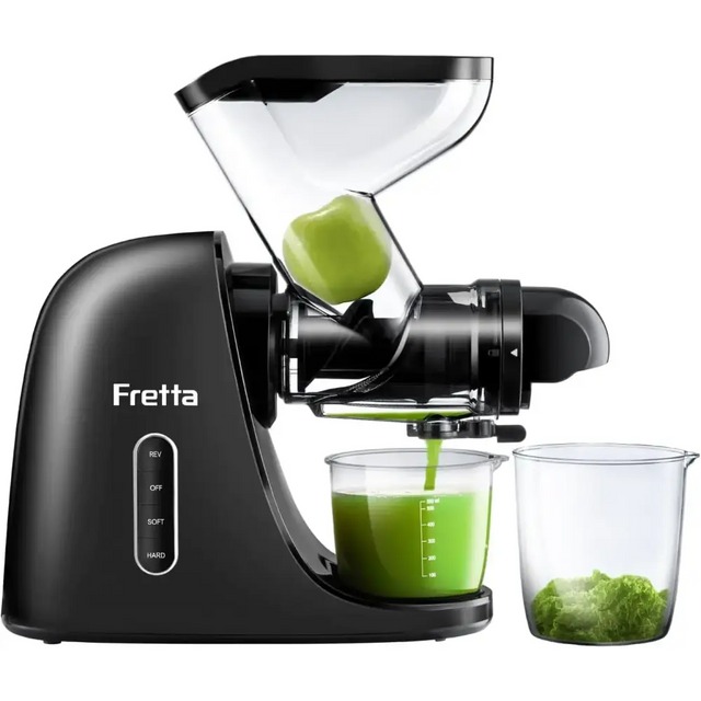 Black fretta juicer with transparent feed chute and clear collection cups, actively extracting green juice from a fruit.