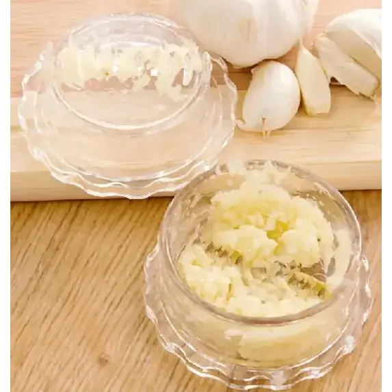 Clear glass garlic press with scalloped edges and a matching lid, containing finely minced garlic.