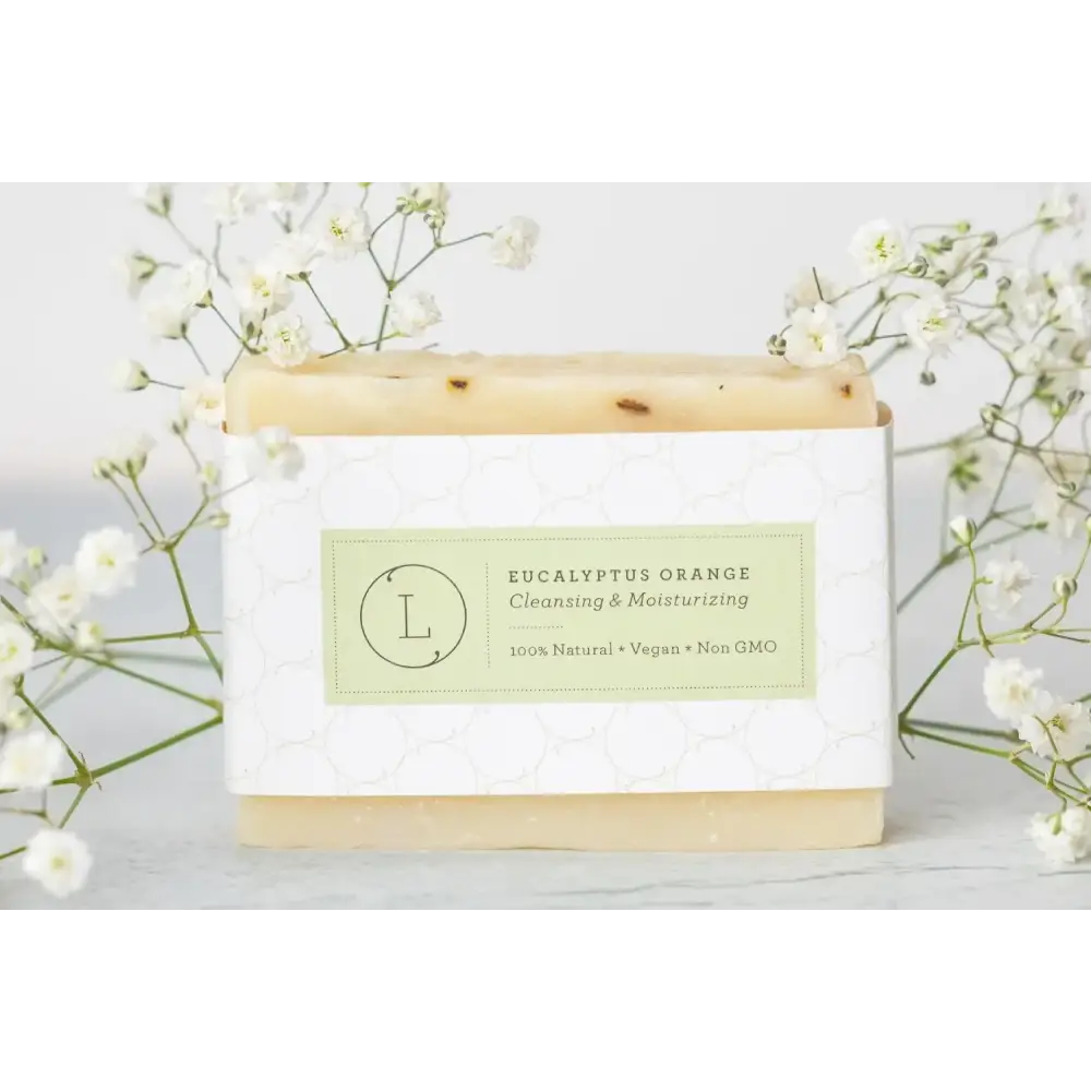 A rectangular bar of pale beige eucalyptus orange soap with visible speckles, wrapped in a white patterned paper label featuring green text and an ’l’ logo.