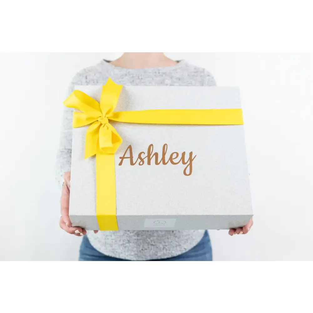White gift box with a bright yellow ribbon bow and the name ’ashley’ in gold cursive lettering.