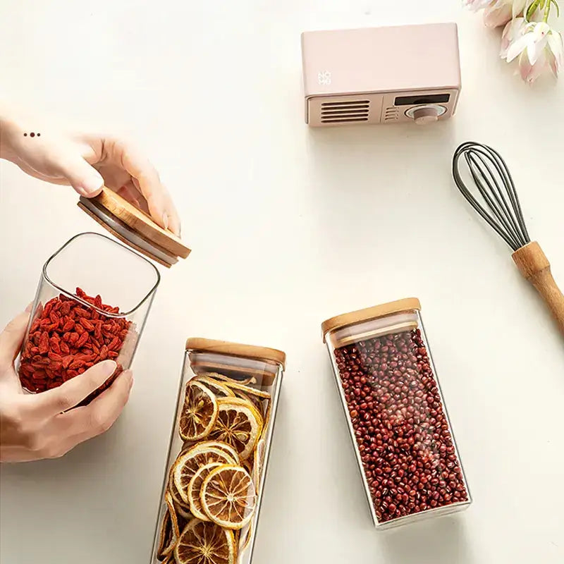 Clear glass rectangular storage jars with bamboo lids, filled with dried goji berries, citrus slices, and red beans.