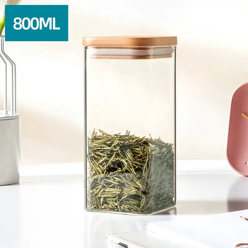 Clear glass storage jar with a light wood lid, filled with green tea leaves.