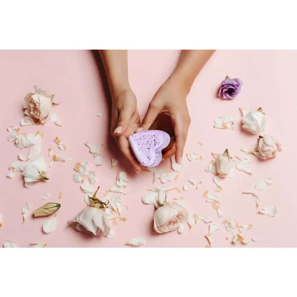 A lavender heart-shaped bath bomb with a speckled texture, held gently in two hands against a soft pink background scattered with rose petals and flowers.