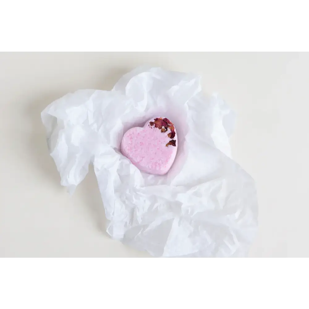 A pink heart-shaped bath bomb with dried rose petals embedded in its surface, nestled within crumpled white tissue paper.