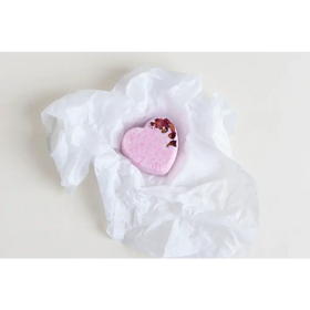 A pink heart-shaped bath bomb with dried rose petals embedded in its surface, nestled within crumpled white tissue paper.