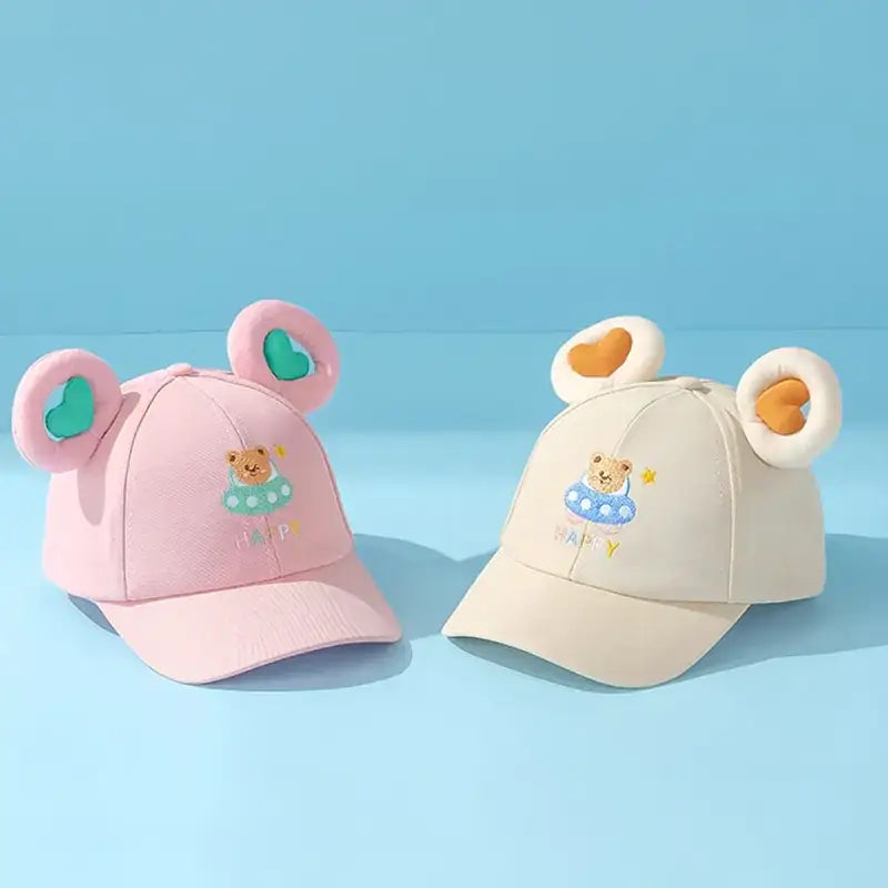 Pink and cream-colored toddler baseball caps featuring plush bear ears with heart-shaped cutouts, each adorned with an embroidered teddy bear in a swimsuit.