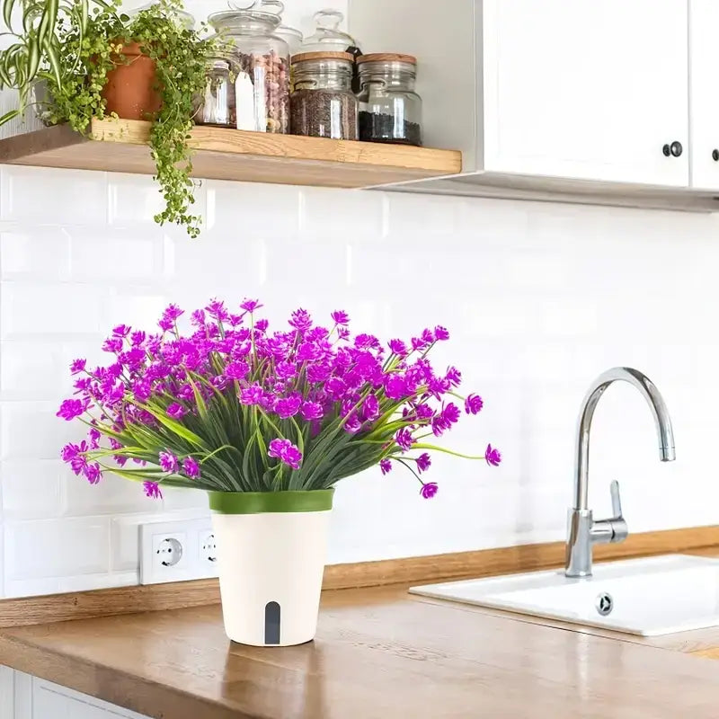 A vibrant purple artificial flower arrangement in a white and green ceramic pot with a small black rectangular detail on the side.