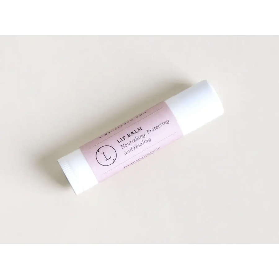 A pale pink and white lip balm tube with a minimalist label featuring the text ’lip balm nourishing, protecting and healing’ and a circular logo containing an ’l’.