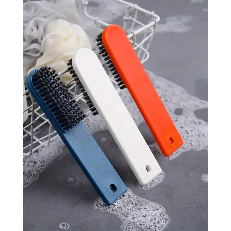 Three colorful, plastic-handled scrub brushes with dense black bristles—blue, white, and orange—are arranged in a wire basket beside soapy water.