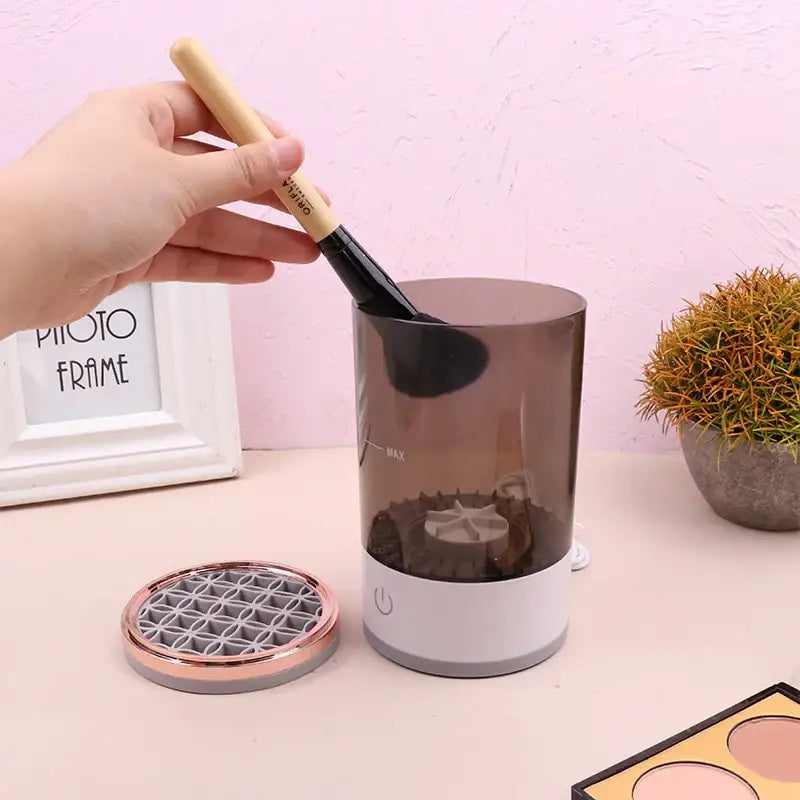 Transparent brown cylindrical makeup brush cleaner with white base and power button, featuring a rose gold mesh lid nearby