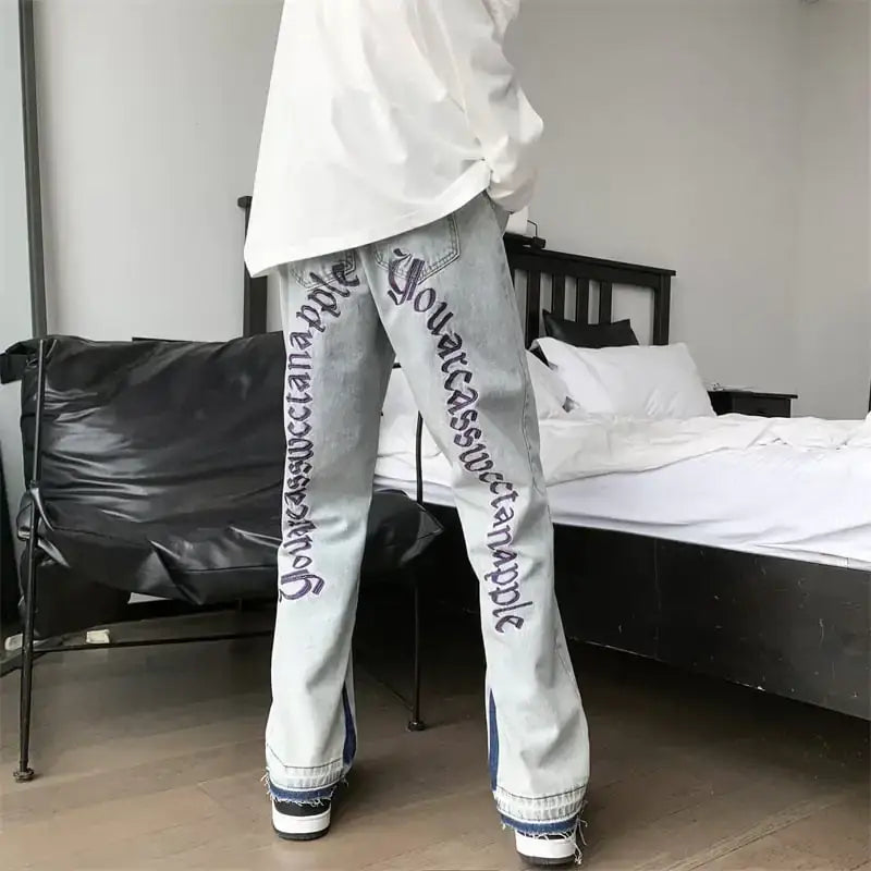 Light-wash denim jeans featuring bold purple script lettering down the outer leg and frayed, multi-colored hem detailing.