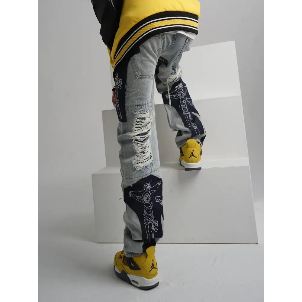Distressed light-wash denim jeans featuring bold black panels with white line-art crucifixion designs and frayed, ripped details.