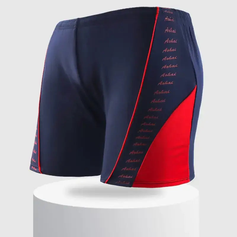 Navy blue and red swim trunks with a bold diagonal color block design, featuring the word ’asha’ printed in pink along the side seam.